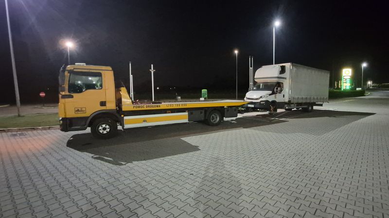 Żółta laweta Pomocy Drogowej Prusakowski holuje nocą biały samochód dostawczy
